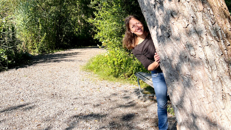 Jeannine Maurer lacht hinter einem Baumstamm hervor. Pause beim Aufnehmen einer Atemmeditation gegen Stress.