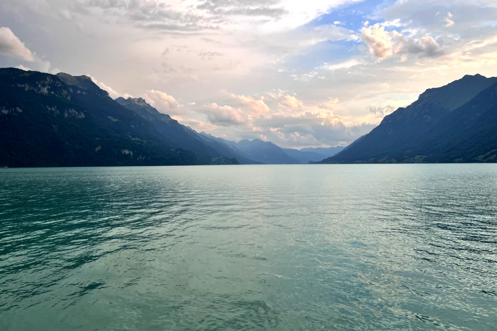 Weitblick vom grünlich scheinenden See in die Berge mit bewölktem Himmel